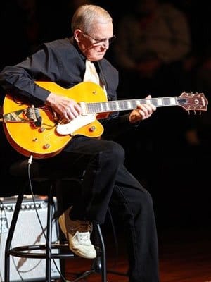 Paul Yandell, CGP, sitting on a stool on stage playing guitar